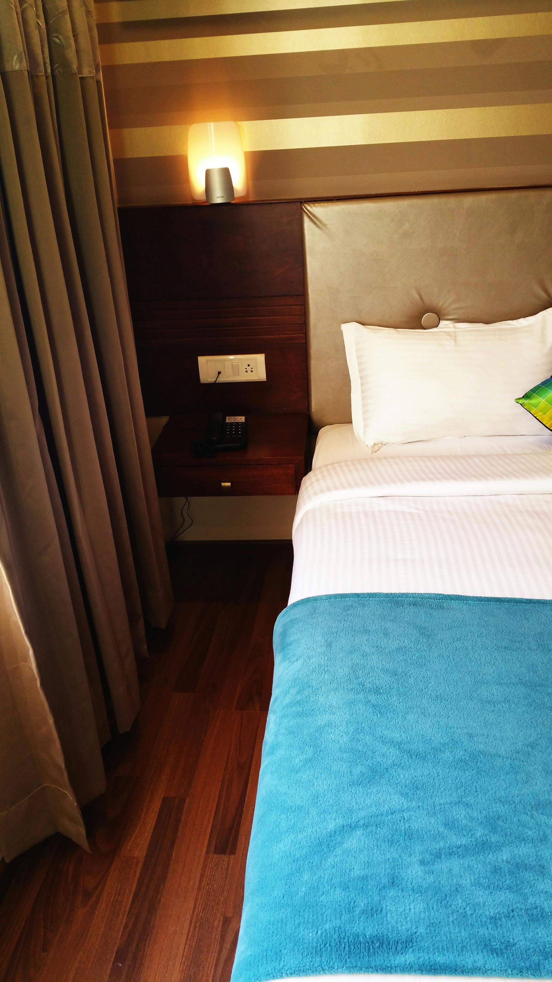 A hotel bedroom with a bed featuring white linens and a turquoise throw, next to a dark wood nightstand and lamp.