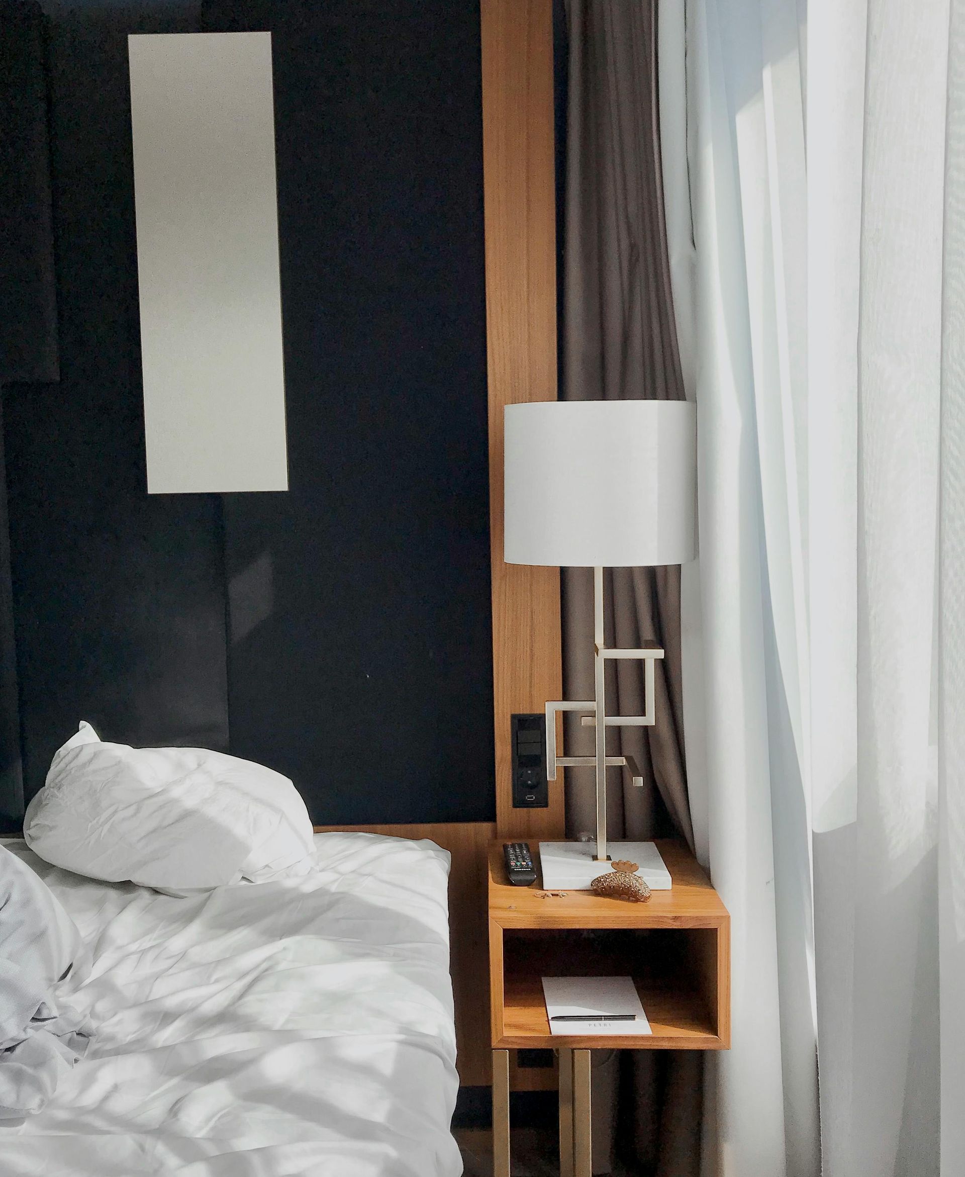 A bedside table with a modern geometric lamp and a book next to a white unmade bed, set against a dark wall and curtains.