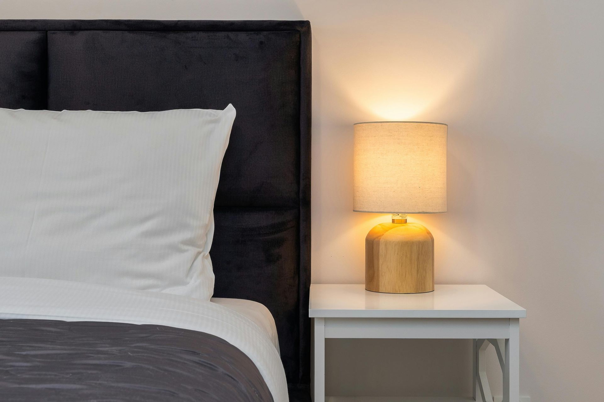 A dark upholstered headboard with a white pillow and grey bedding, next to a white nightstand with a wooden lamp.