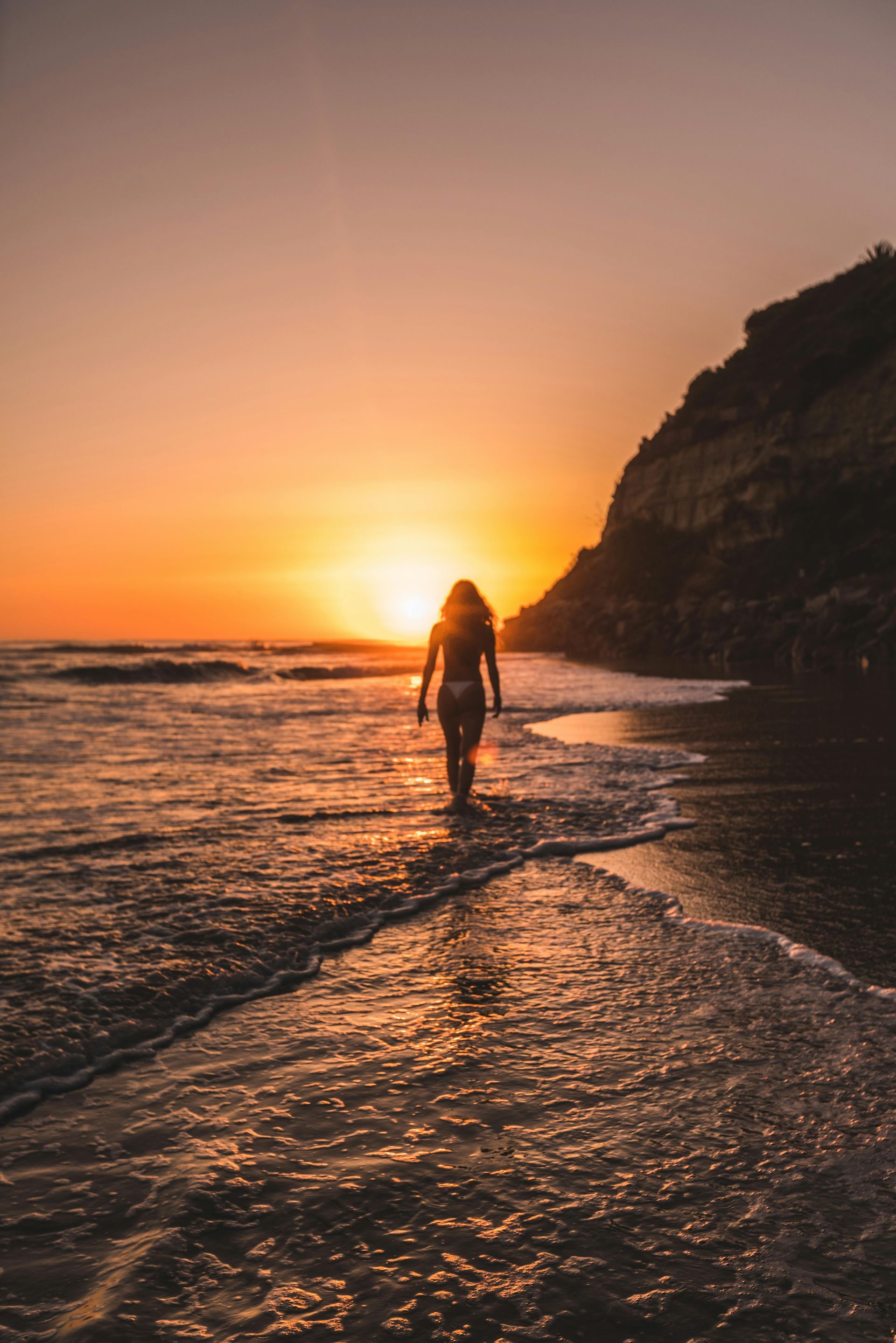 A person walking along the water's edge on a beach at sunset, with a cliffside in the background.