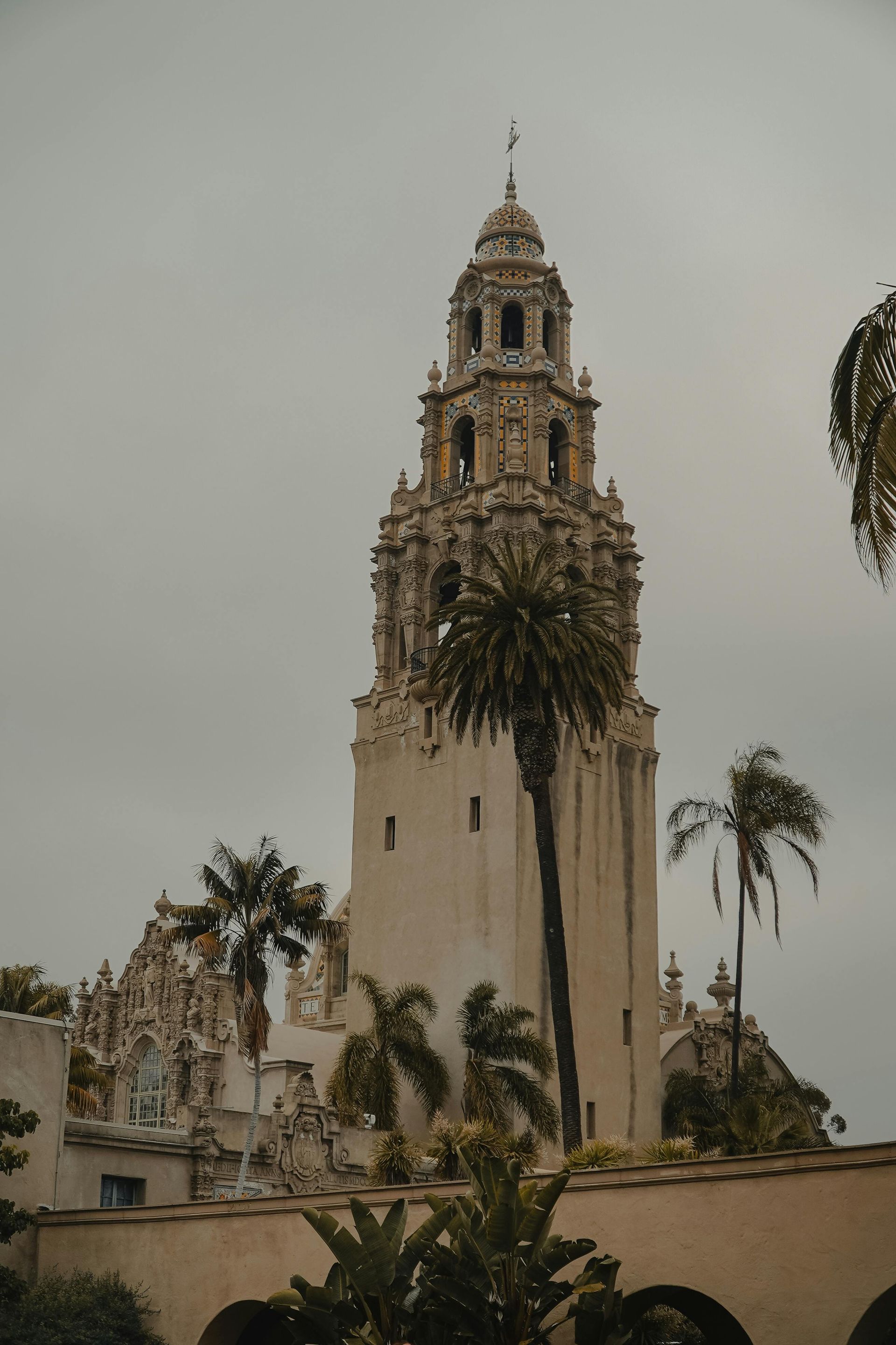 An ornate tower with Spanish-style architecture rises above palm trees against a cloudy, muted sky.