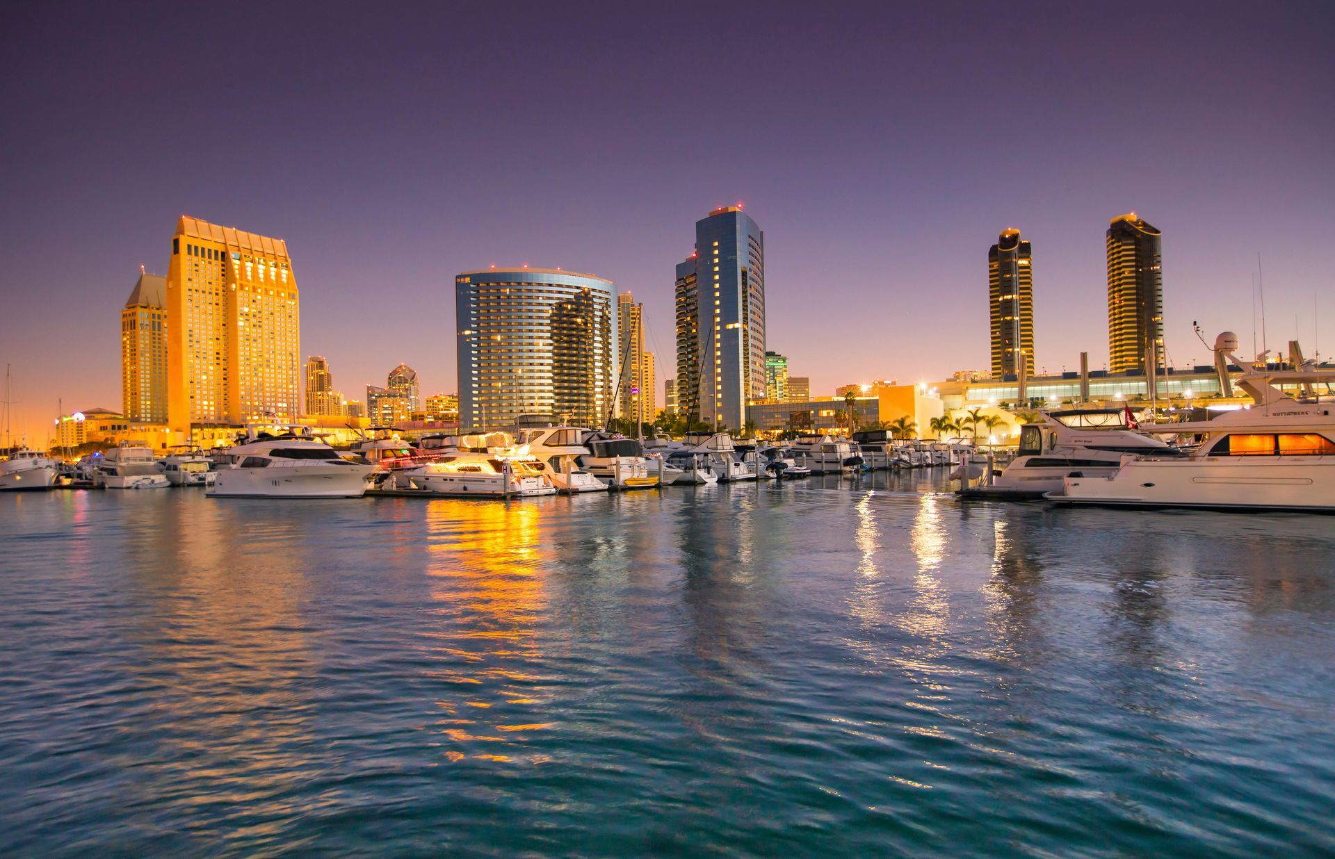 San Diego skyline at twilight, featuring illuminated buildings reflecting over a marina filled with yachts.