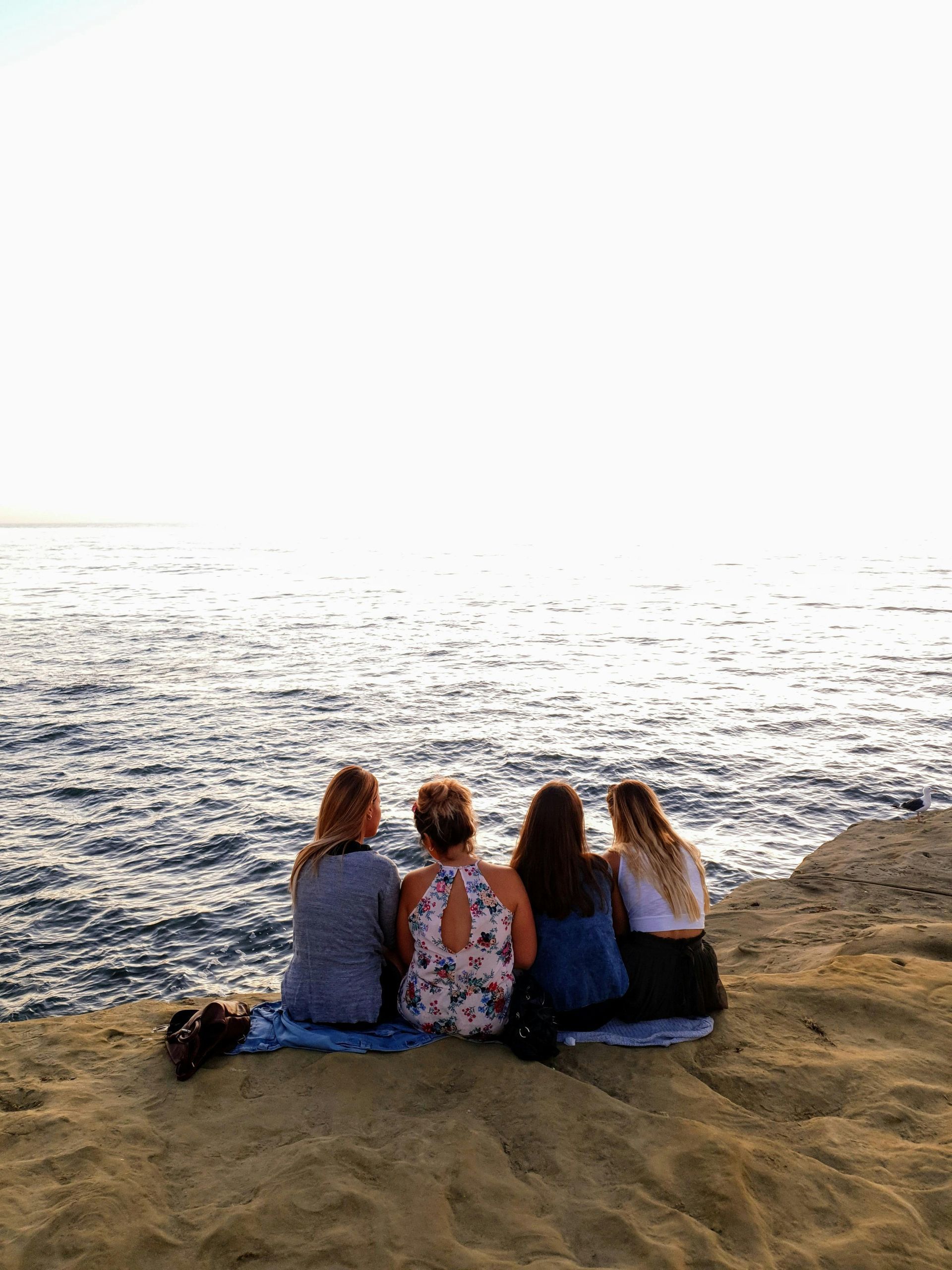 Four people sitting together on a rocky cliff edge, overlooking the sea during sunset.