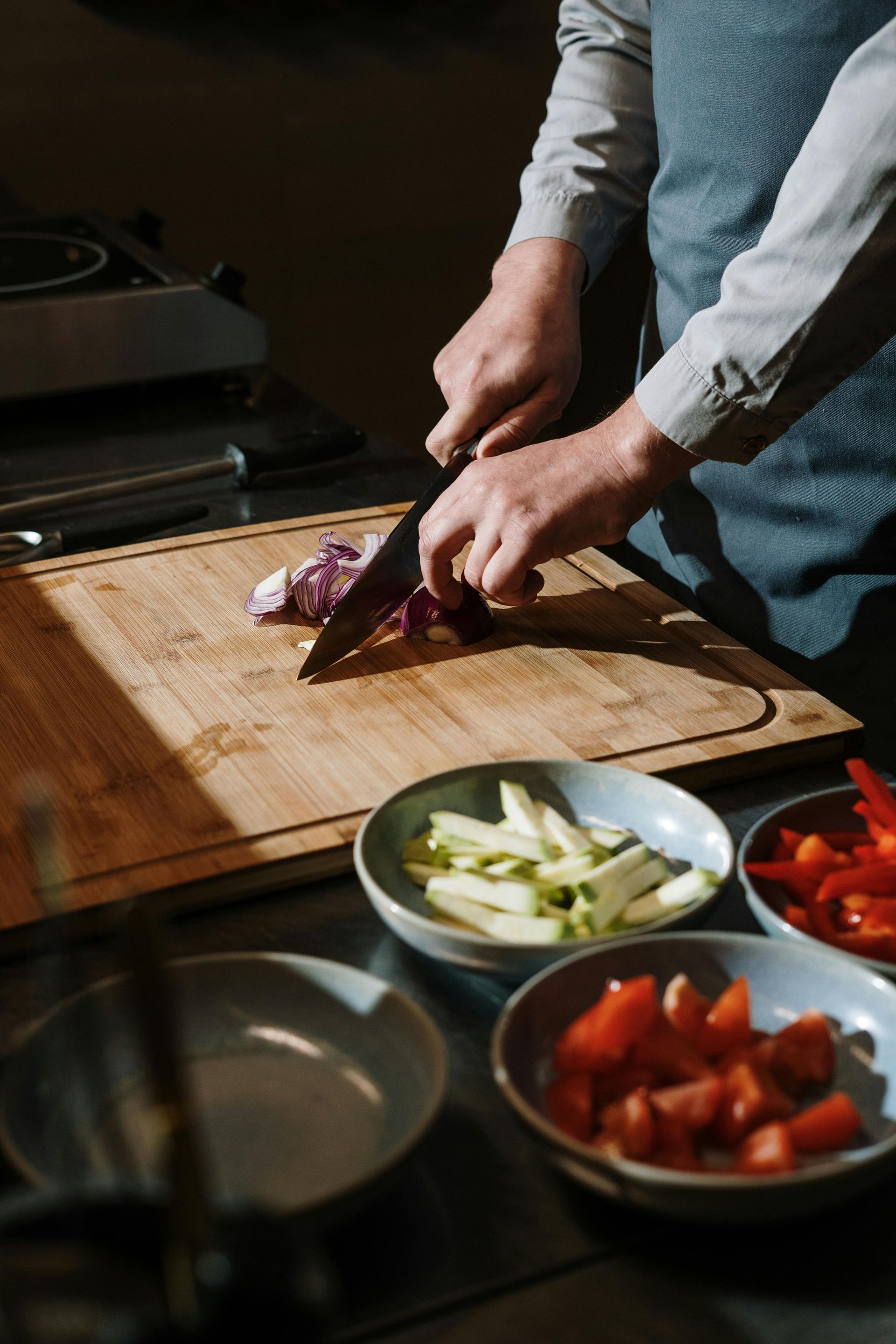 A person in a blue apron chops a red onion on a wooden cutting board, with bowls of sliced vegetables nearby.