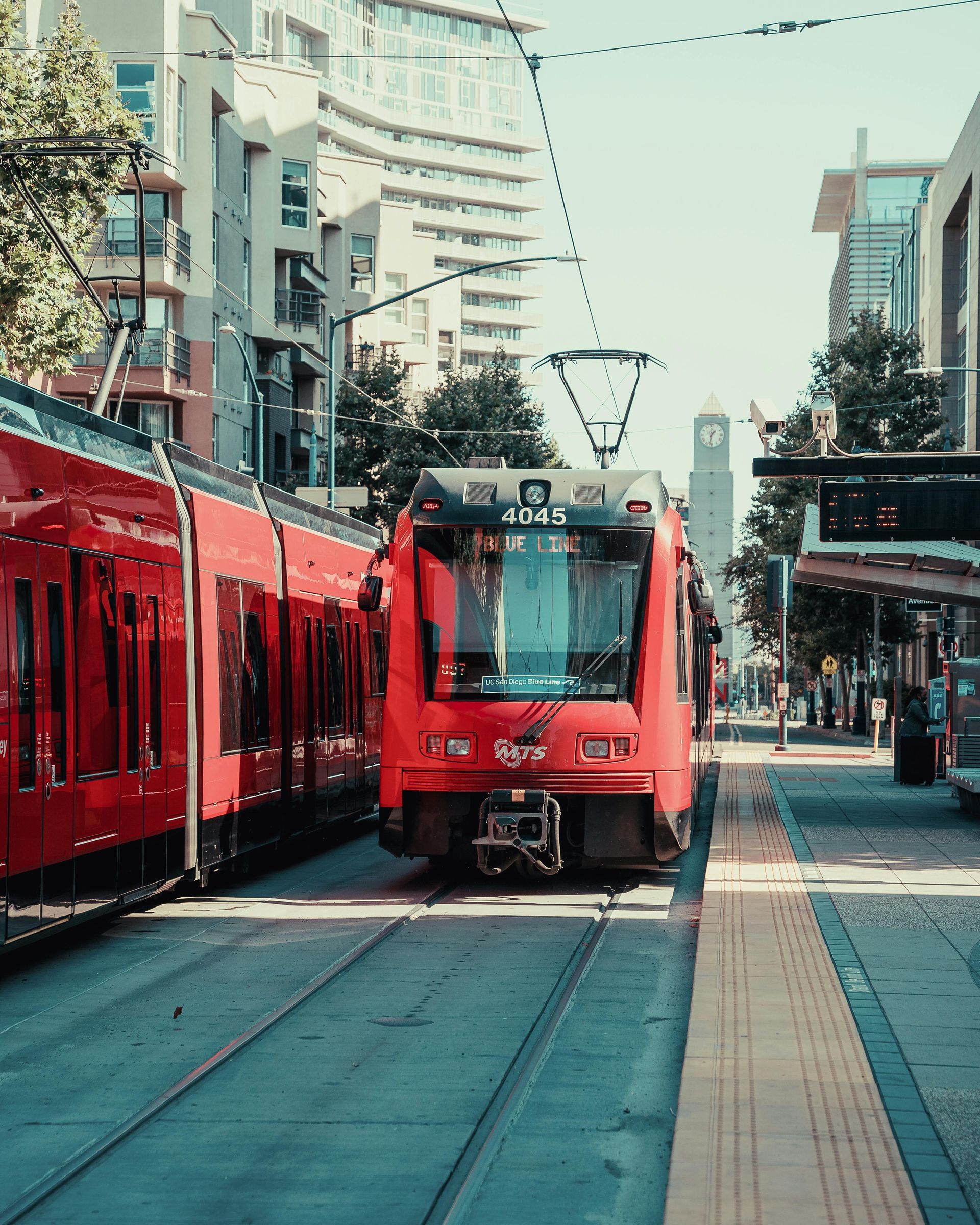 A bright red tram moves along city street tracks past tall buildings on a sunny day.