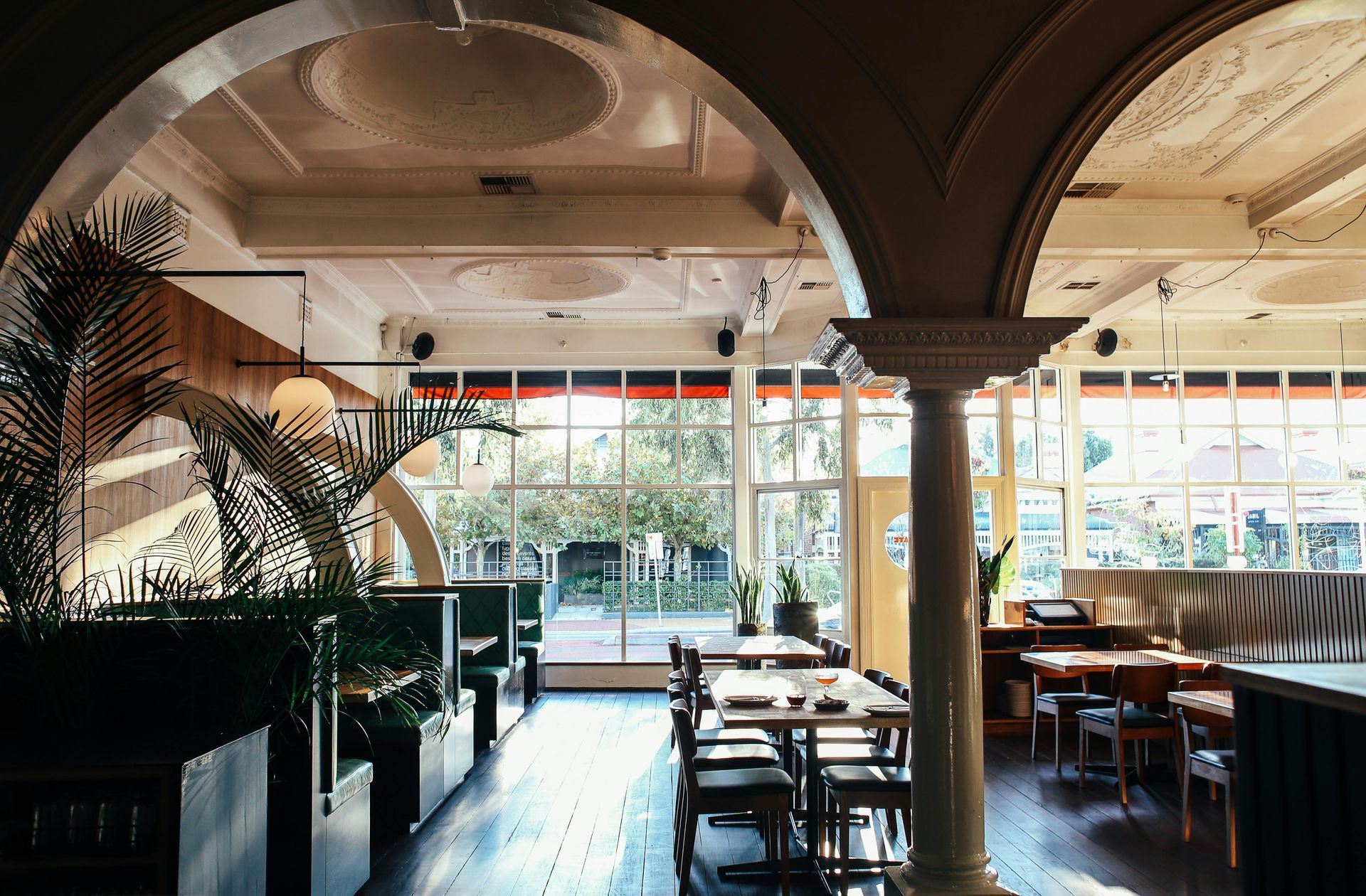 Interior view of a restaurant with arched pillars, dining tables, chairs, and large windows with greenery outside.