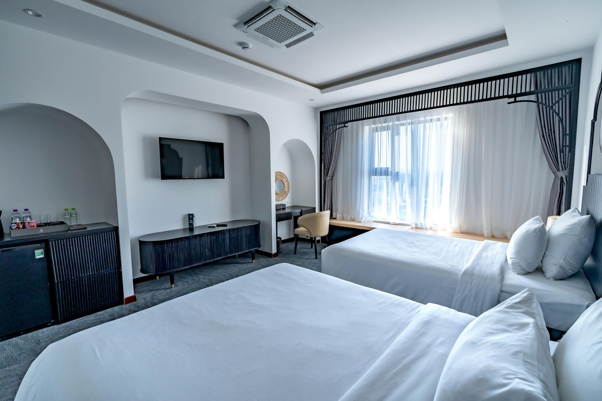 A modern hotel room with two white-made beds, a television, a long desk, and a window with sheer curtains.