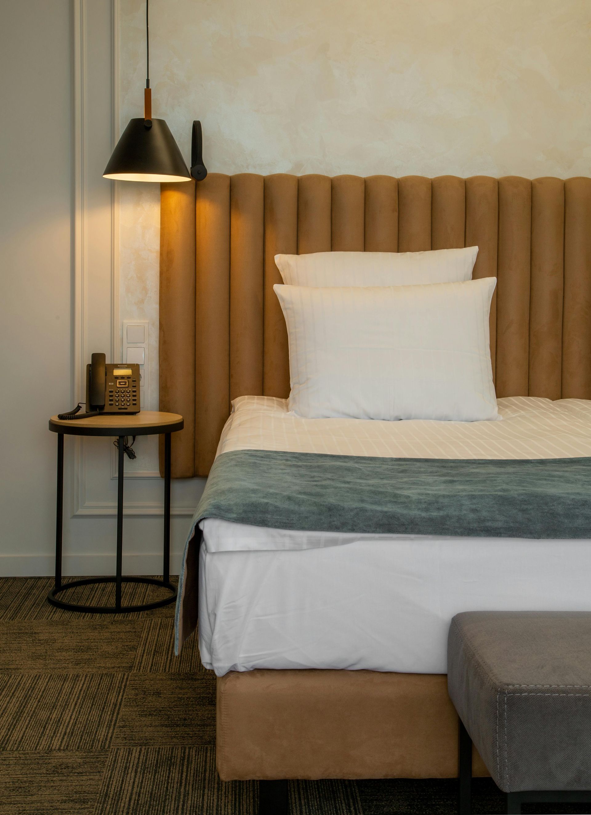 A bed with white linens and a blue runner, against a tan vertical-paneled headboard, next to a black pendant lamp and stand.