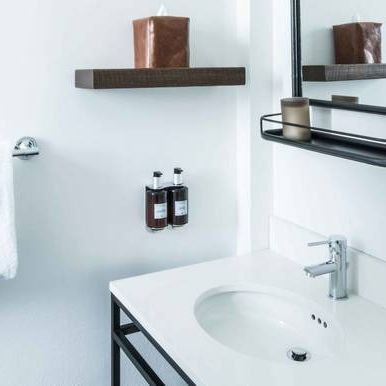 A modern bathroom sink vanity with a white counter, black metal legs, wall-mounted soap dispensers, and wooden shelving.
