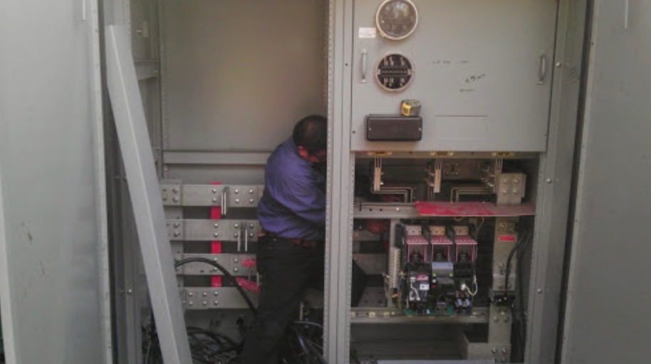Electrician working inside an open gray electrical panel cabinet, likely outdoor, with wires and components visible.