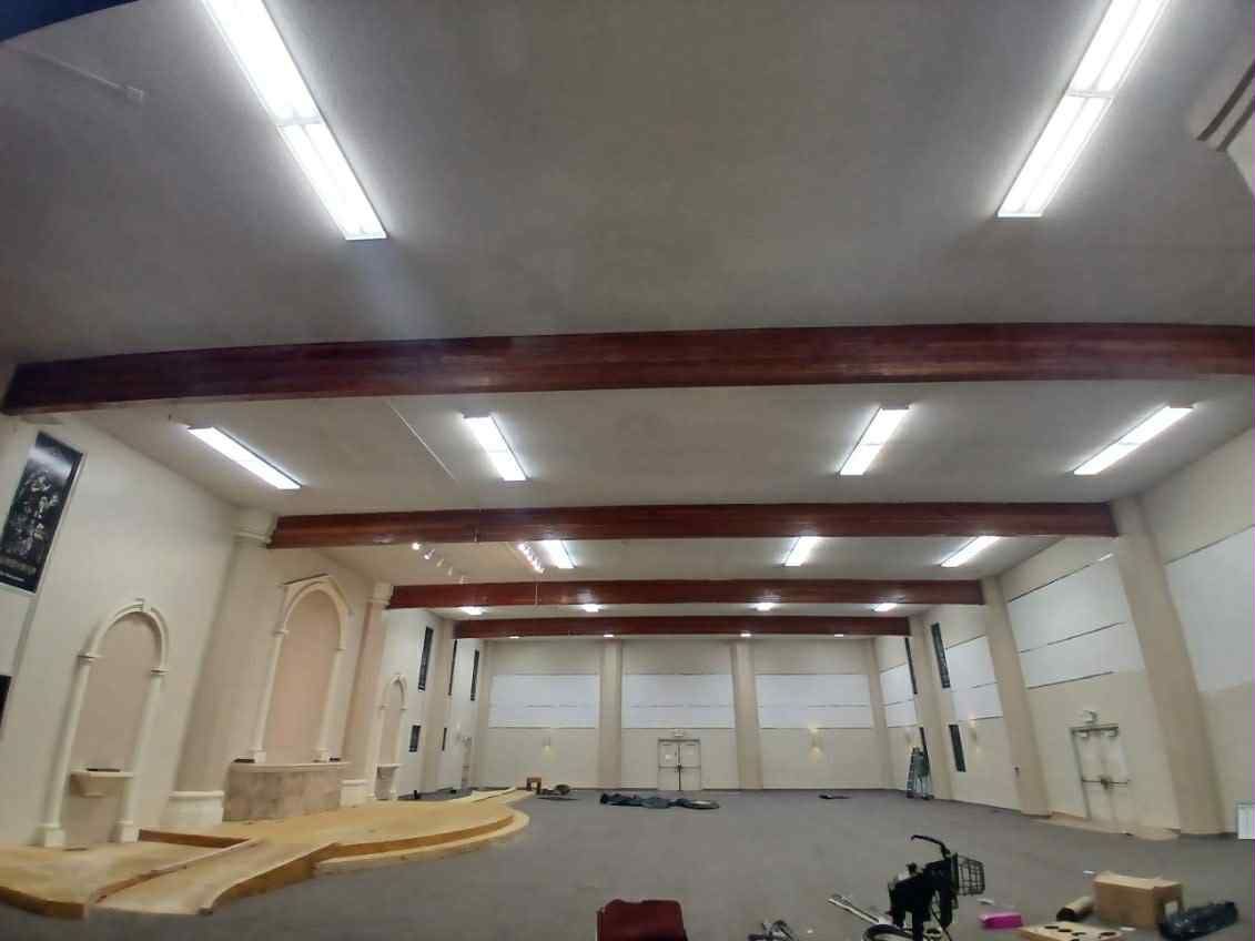 Interior of a building with beige walls, brown beams, and fluorescent lights. A stage is in the foreground.