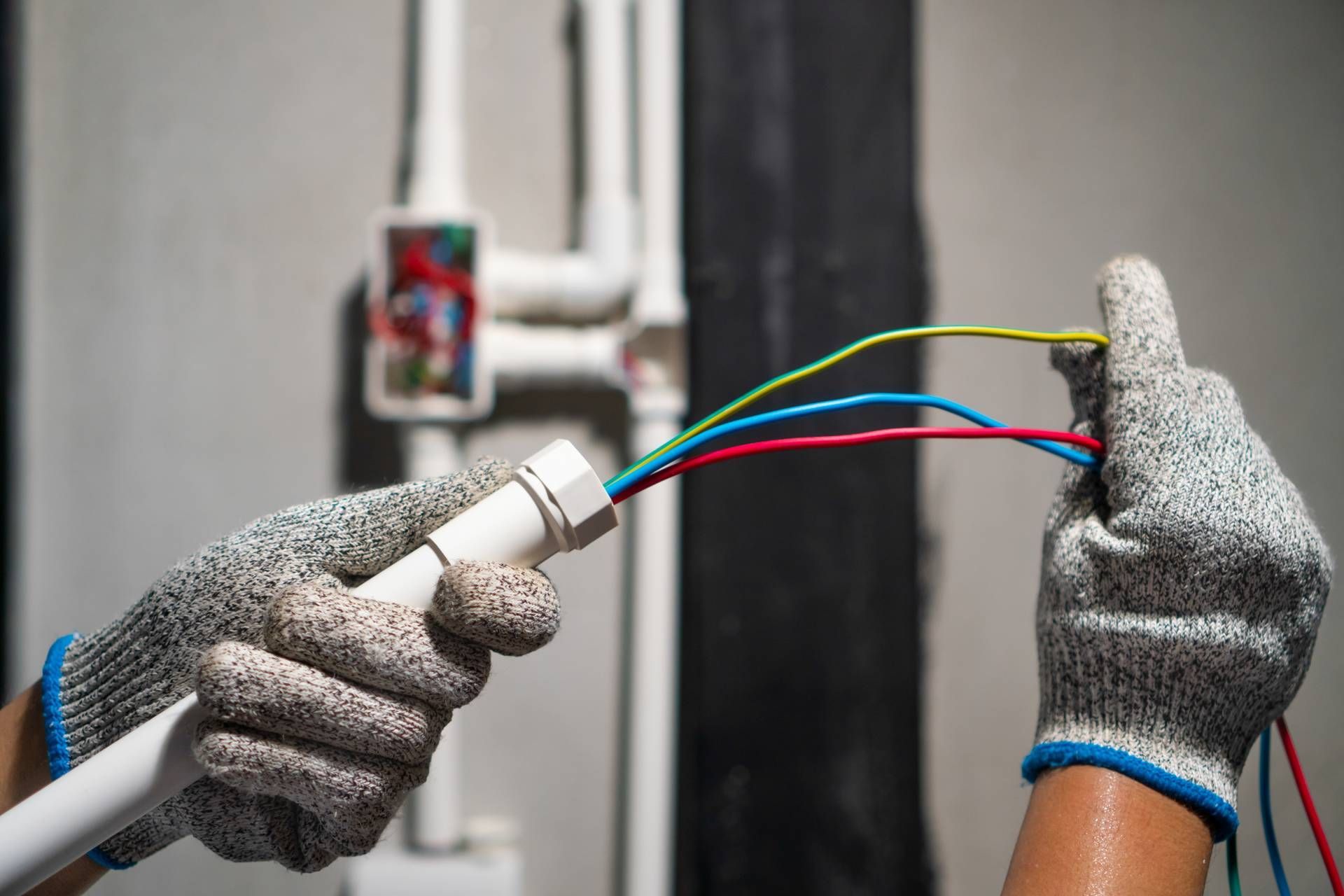 Hands wearing gloves holding wires and a pipe. Electrical work being done indoors.