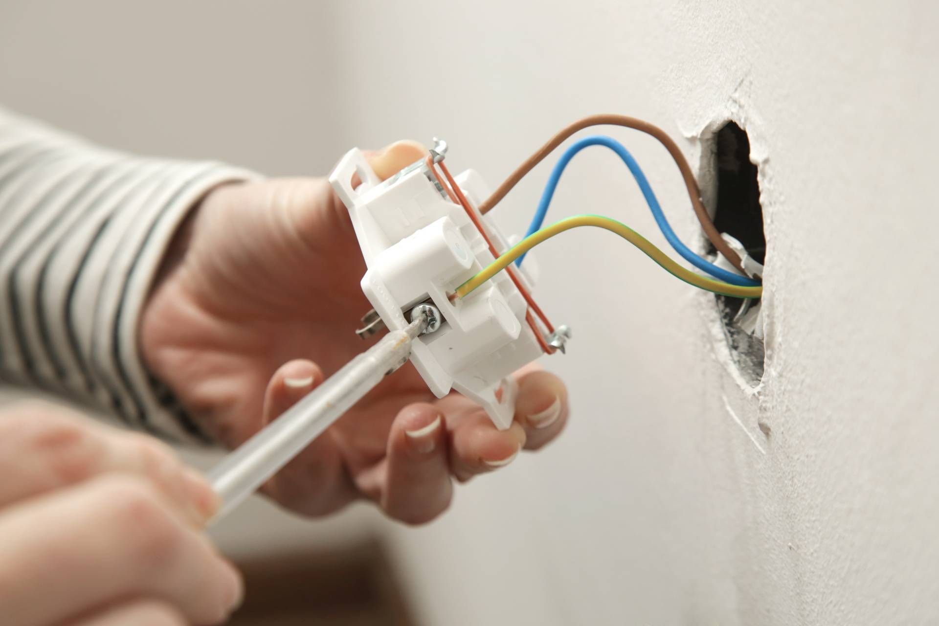 Person installing an electrical outlet, brown, blue, and yellow wires visible. White wall and outlet.