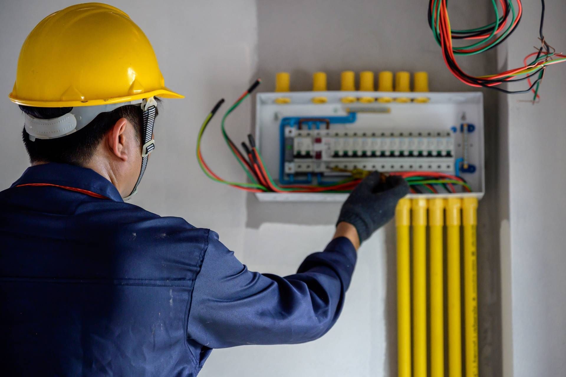 Electrician in blue uniform, yellow hard hat, and gloves, working on a circuit breaker panel.