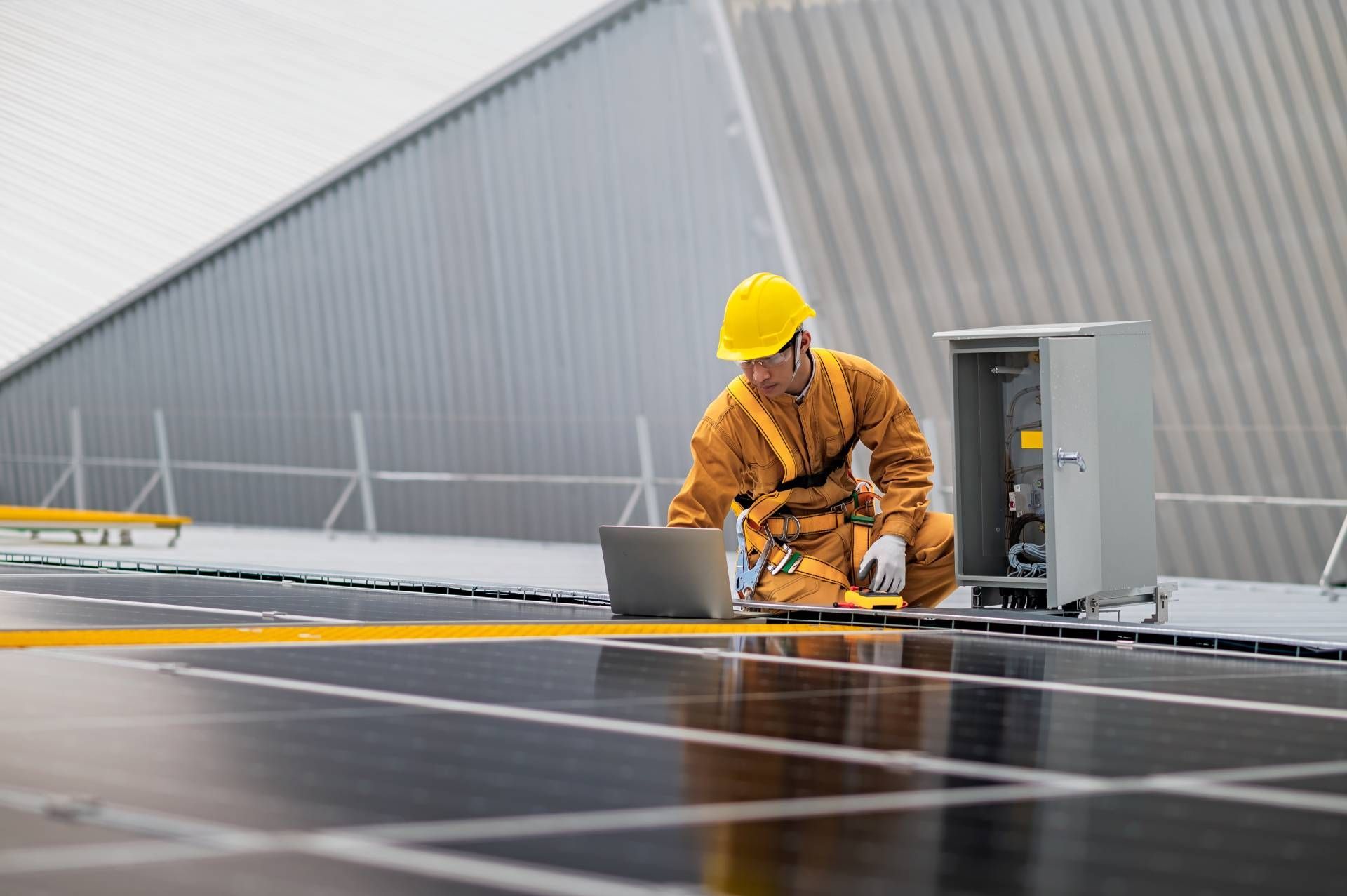 Solar panel technician in yellow hard hat and safety gear working on panels with a laptop on a rooftop.