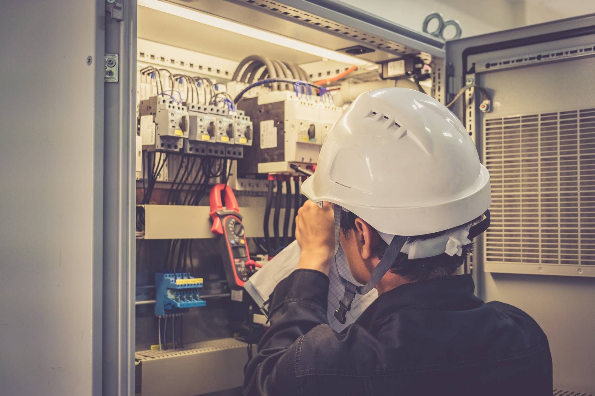 Electrician in a white hard hat reviewing paperwork in front of an open electrical panel.