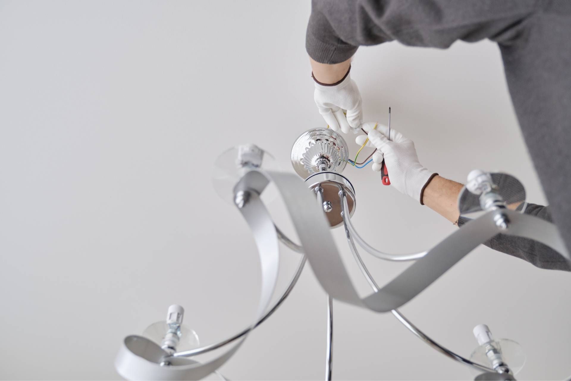 Person in gloves installing a silver chandelier on a white ceiling.