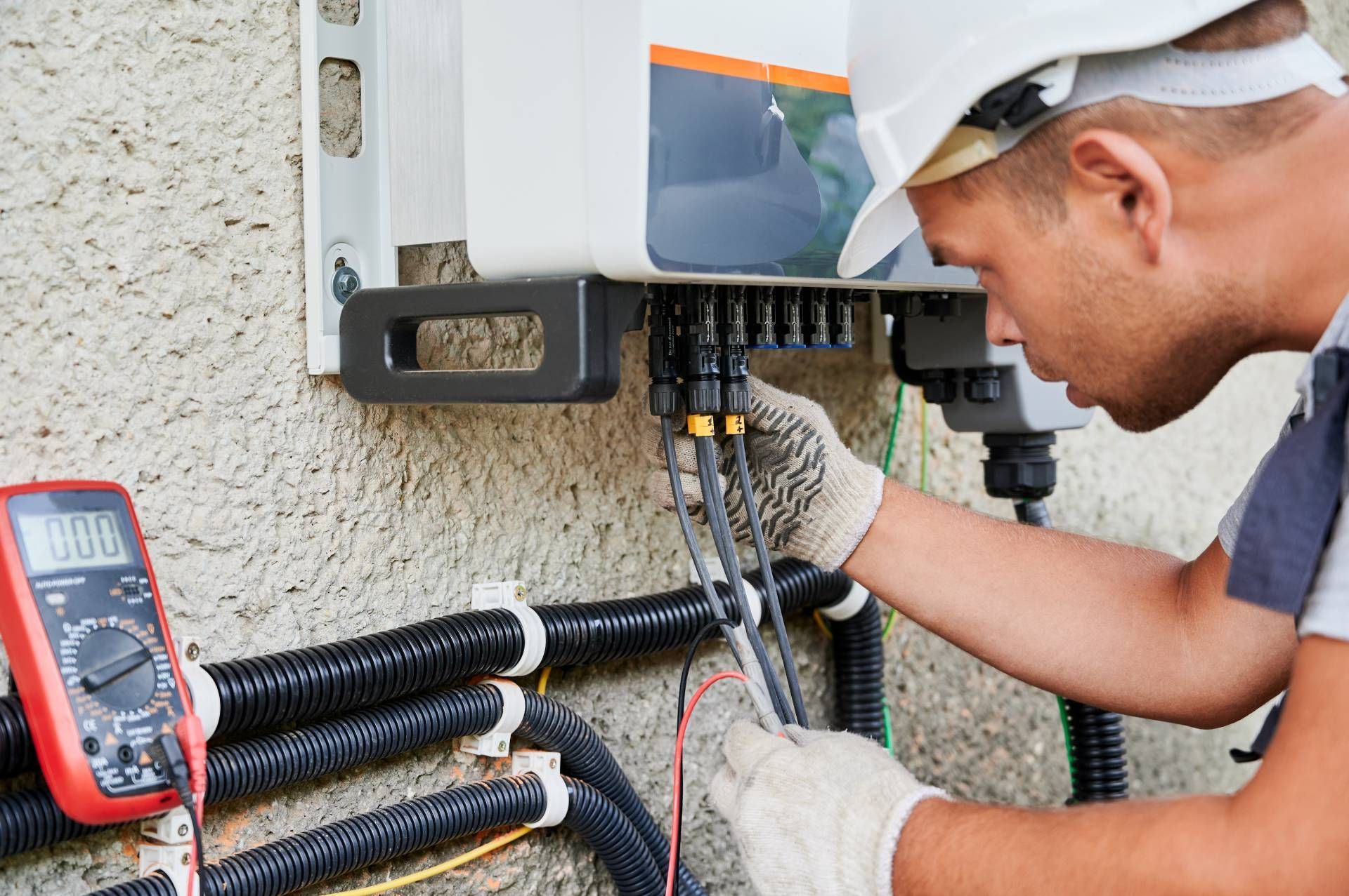Electrician in a hard hat inspecting solar panel wiring with a multimeter on a beige wall.