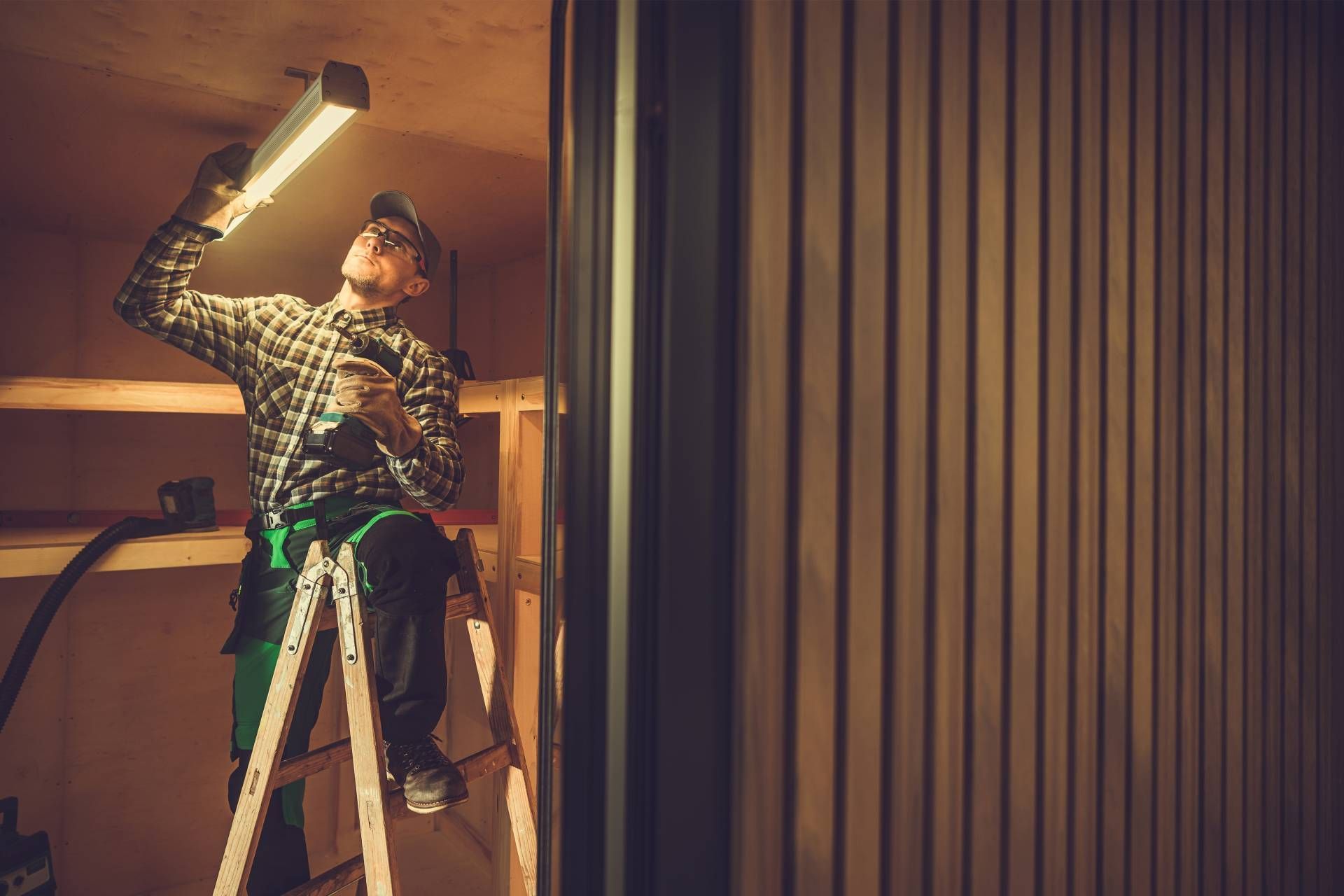 Man on ladder inspecting a light fixture in a dimly lit workshop.