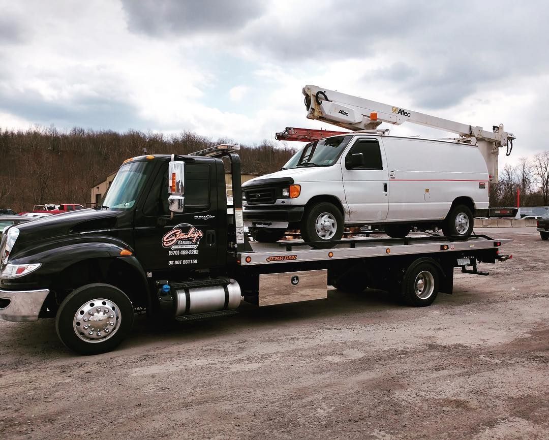 Tow truck carrying a white work van with an aerial lift, on a gravel lot under an overcast sky.