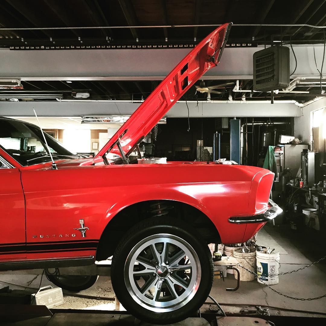 Red 1960s Mustang with hood open in a garage, black racing stripe, chrome wheels.