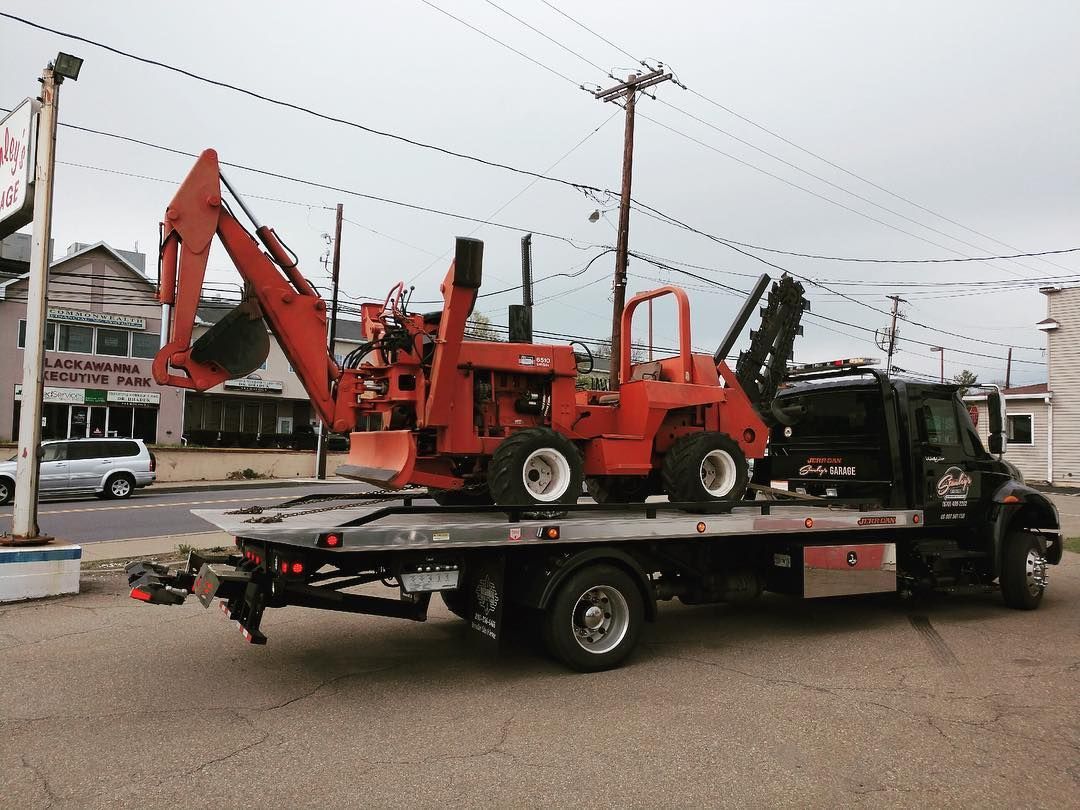 Flatbed tow truck carrying an orange backhoe on a street with buildings and utility poles.