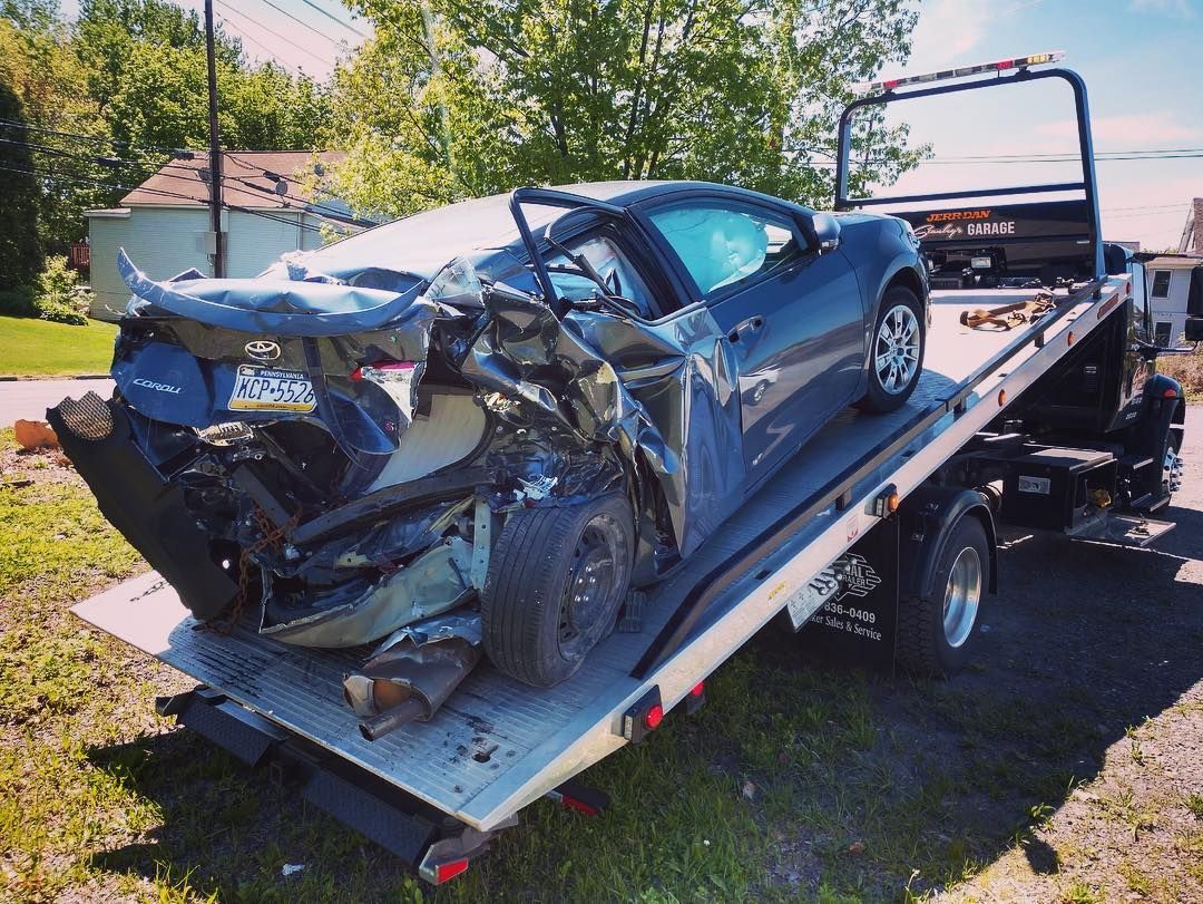 Damaged blue car on a tow truck, rear severely crushed. Outdoor setting, bright sunlight.