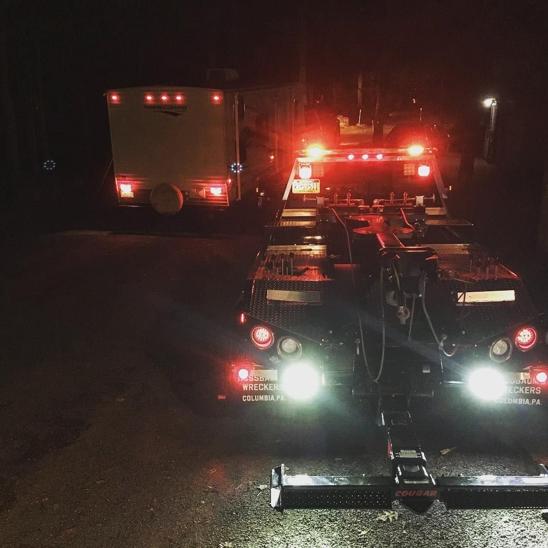 A flatbed tow truck with flashing red lights towing a camper at night.