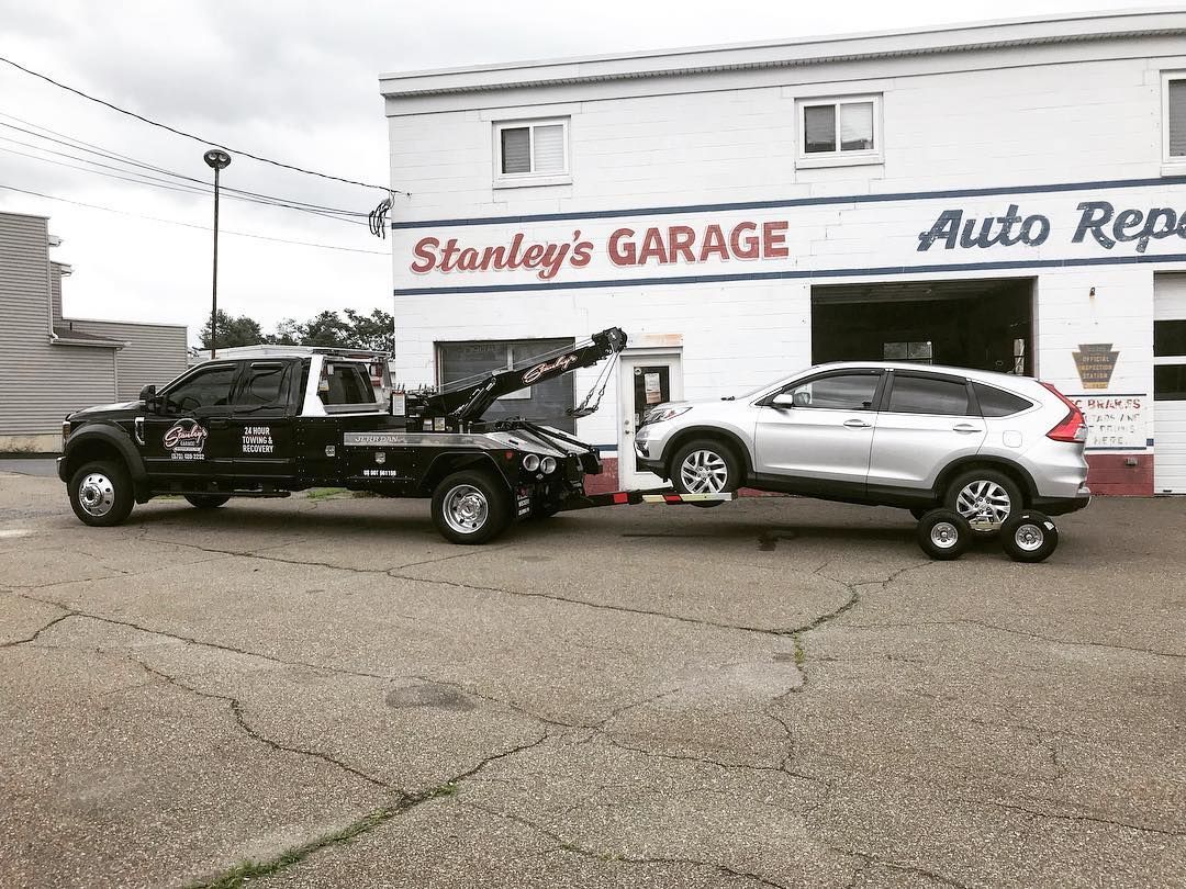 Tow truck towing a silver SUV in front of Stanley's Garage, white building with open garage door.