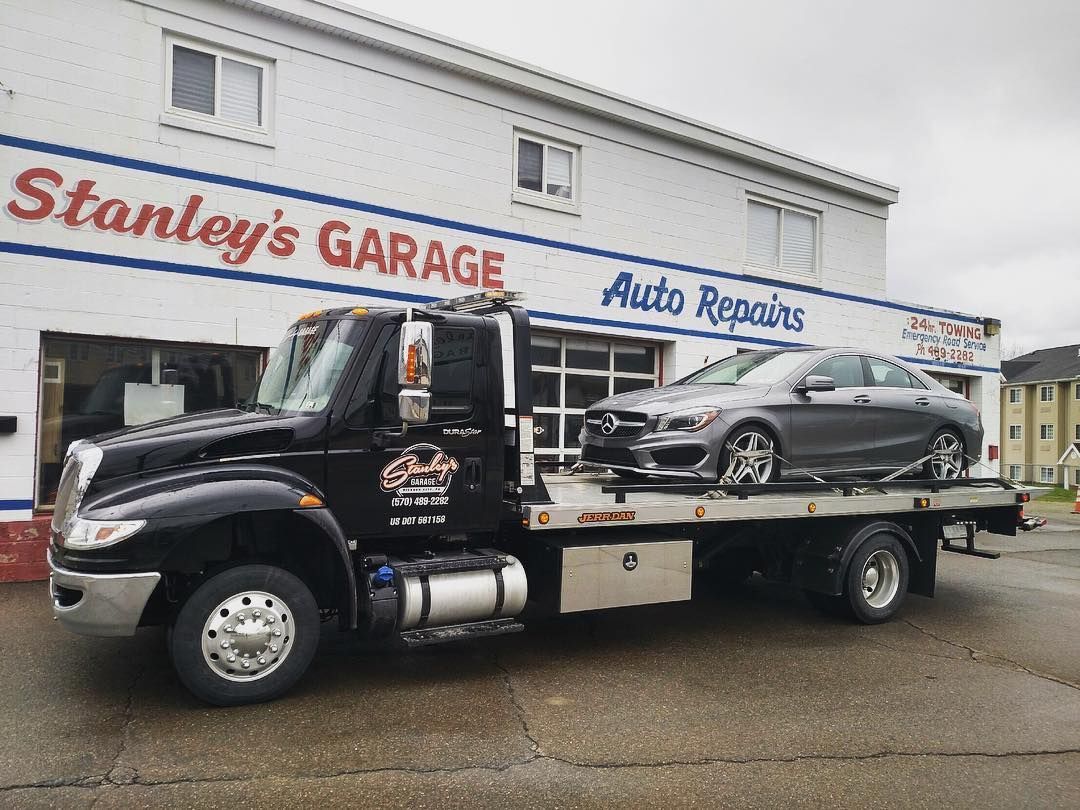 Black tow truck from Stanley's Garage with a gray car on its flatbed, parked in front of the auto repair shop.