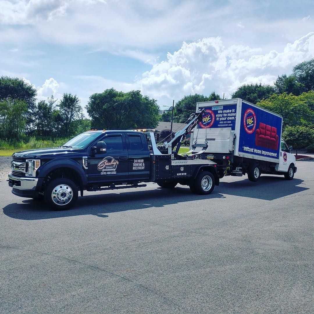 A tow truck towing a box truck on a sunny day. Both trucks have logos.