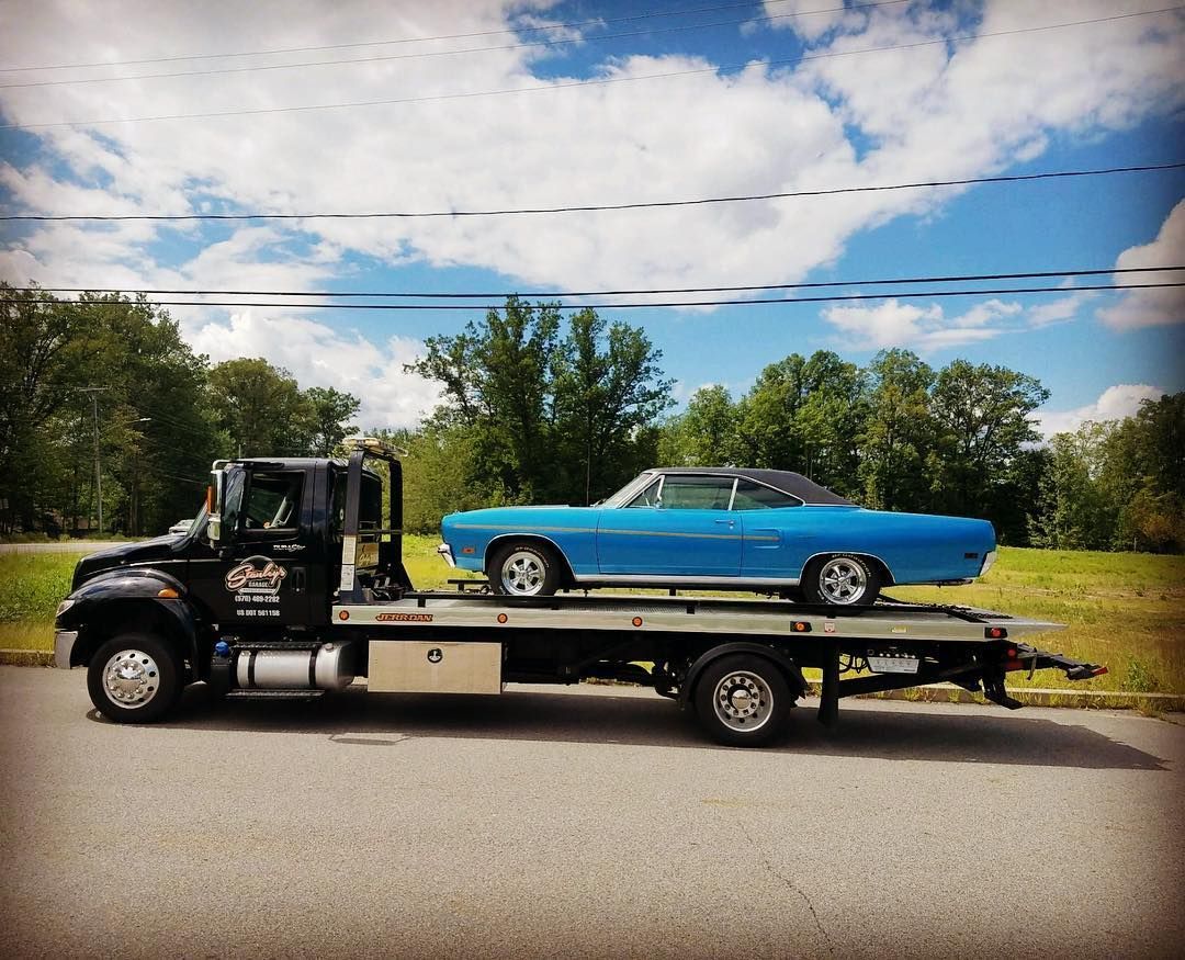 Black tow truck carrying a blue classic car on a sunny day.