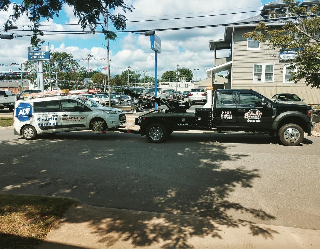 A white van with an ADT logo being towed by a black tow truck on a city street.