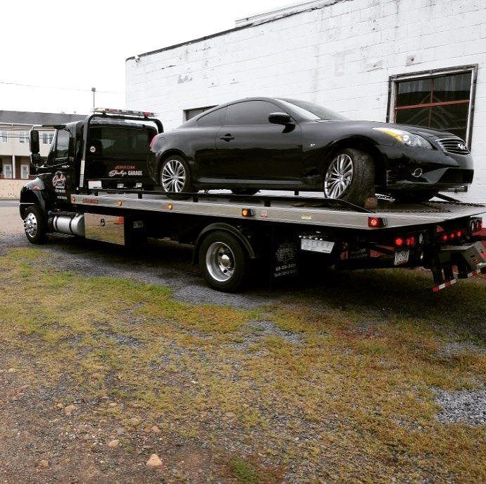 Black car on a flatbed tow truck in front of a white brick building.