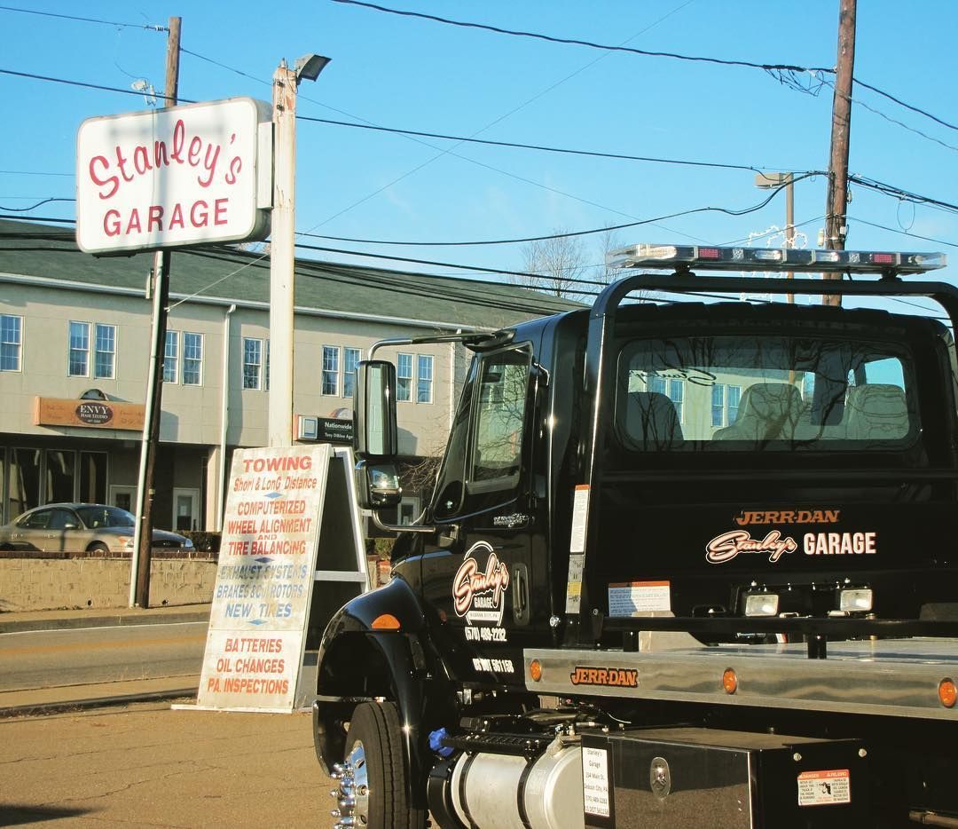 Tow truck parked in front of Stanley's Garage sign. Buildings and power lines in the background on a sunny day.