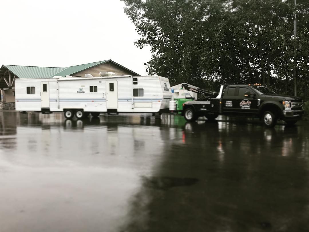 A black tow truck pulling a white camper on a wet, paved surface. A building with a green roof is in the background.