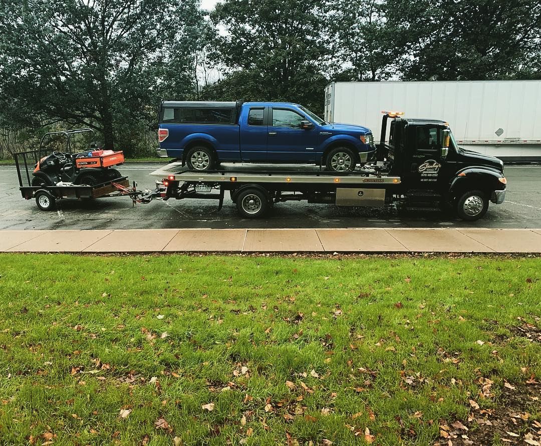 Blue truck on a tow truck, towing a riding lawnmower on a trailer, on a road.