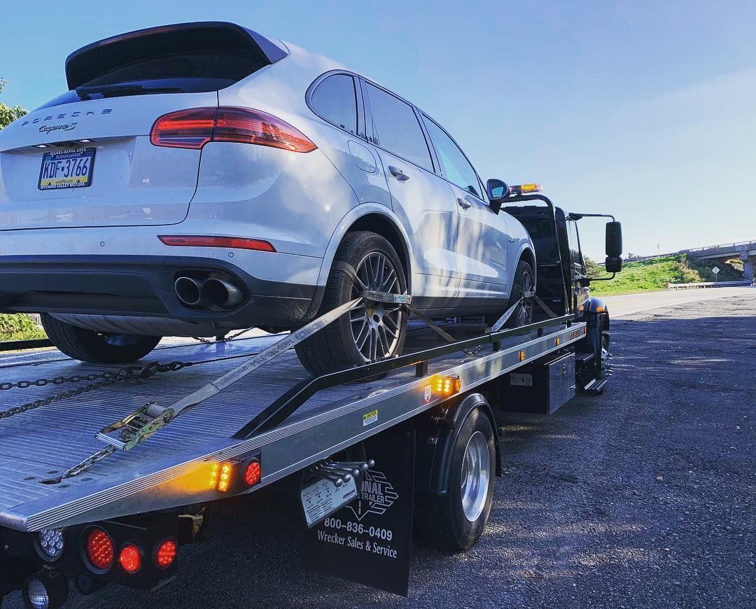 White Porsche Cayenne SUV loaded onto a flatbed tow truck on a sunny day.