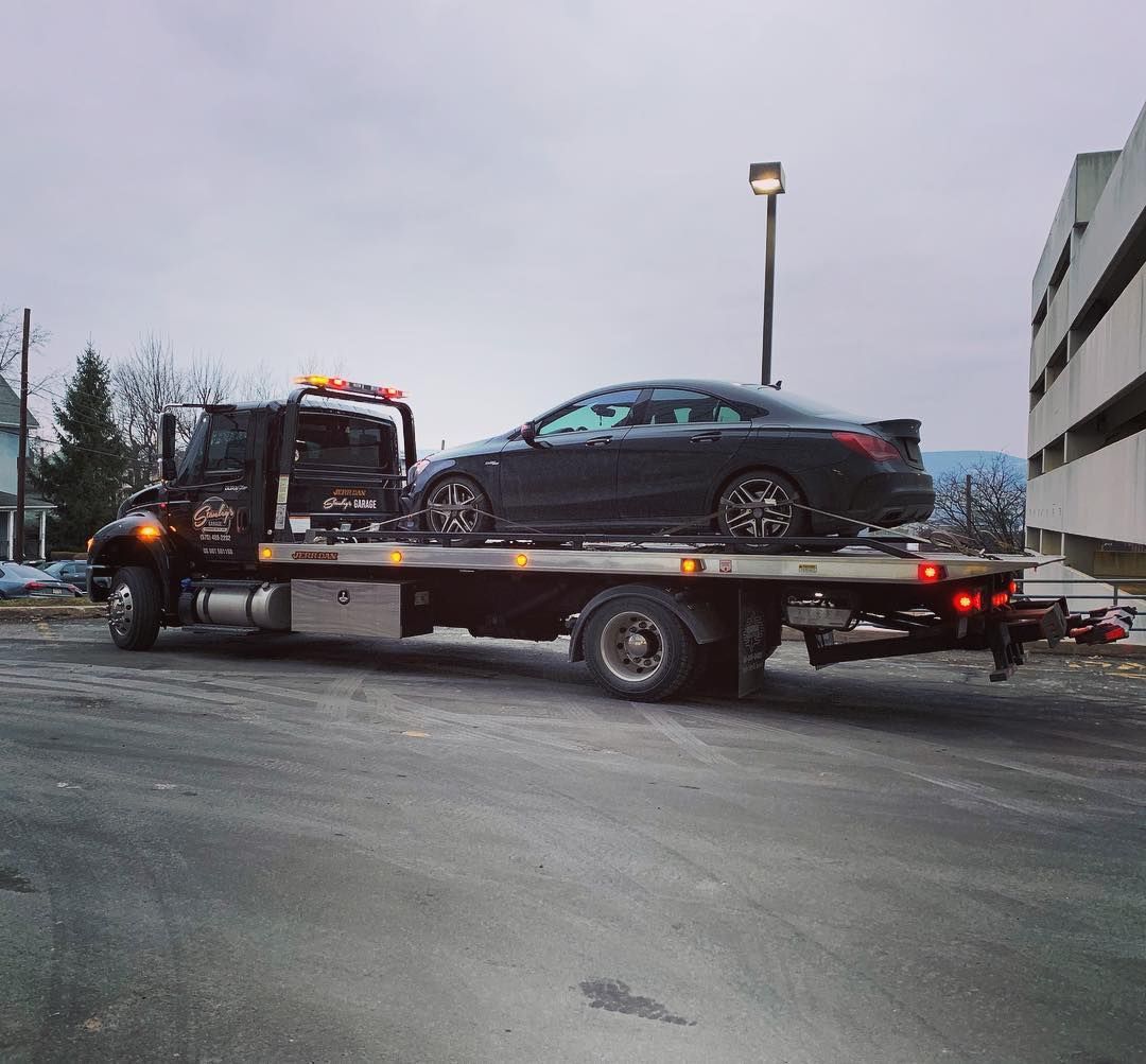 Tow truck carrying a black car on its flatbed in a parking area, overcast sky.