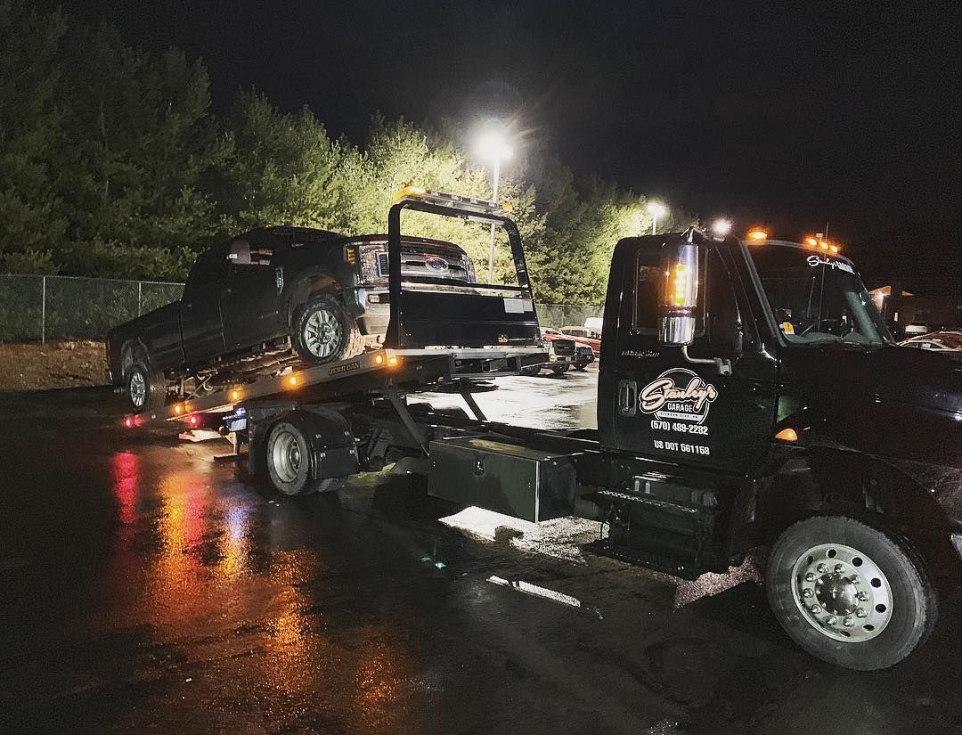 Tow truck carrying a severely burned vehicle at night. Road surface is wet, and lights are illuminated.