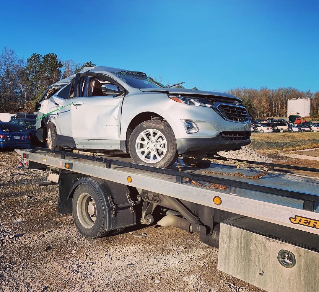 Silver SUV damaged in accident, loaded onto a flatbed tow truck outdoors on a gravel lot.