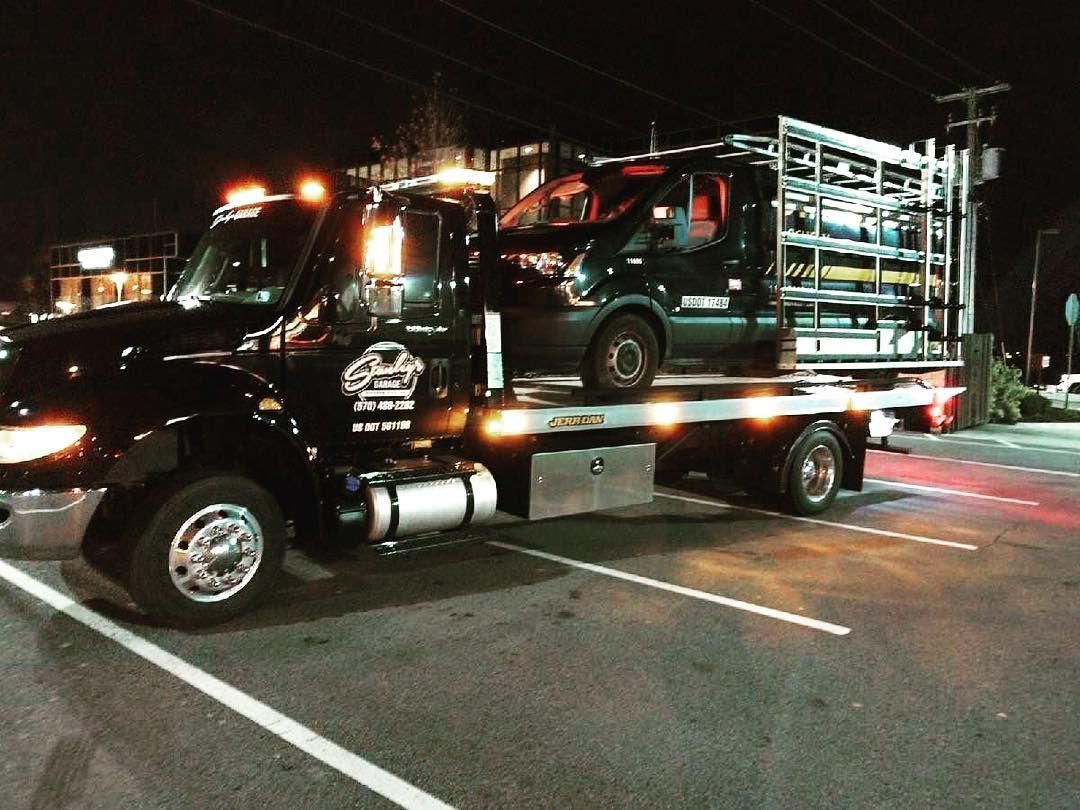 Black tow truck carrying a black van on a flatbed at night, parked near a building.