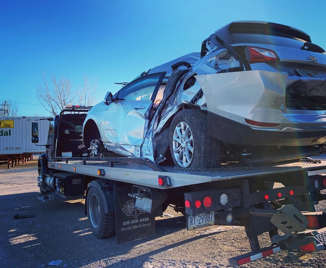 Damaged silver SUV on a tow truck, roadside with blue sky. Significant side damage.