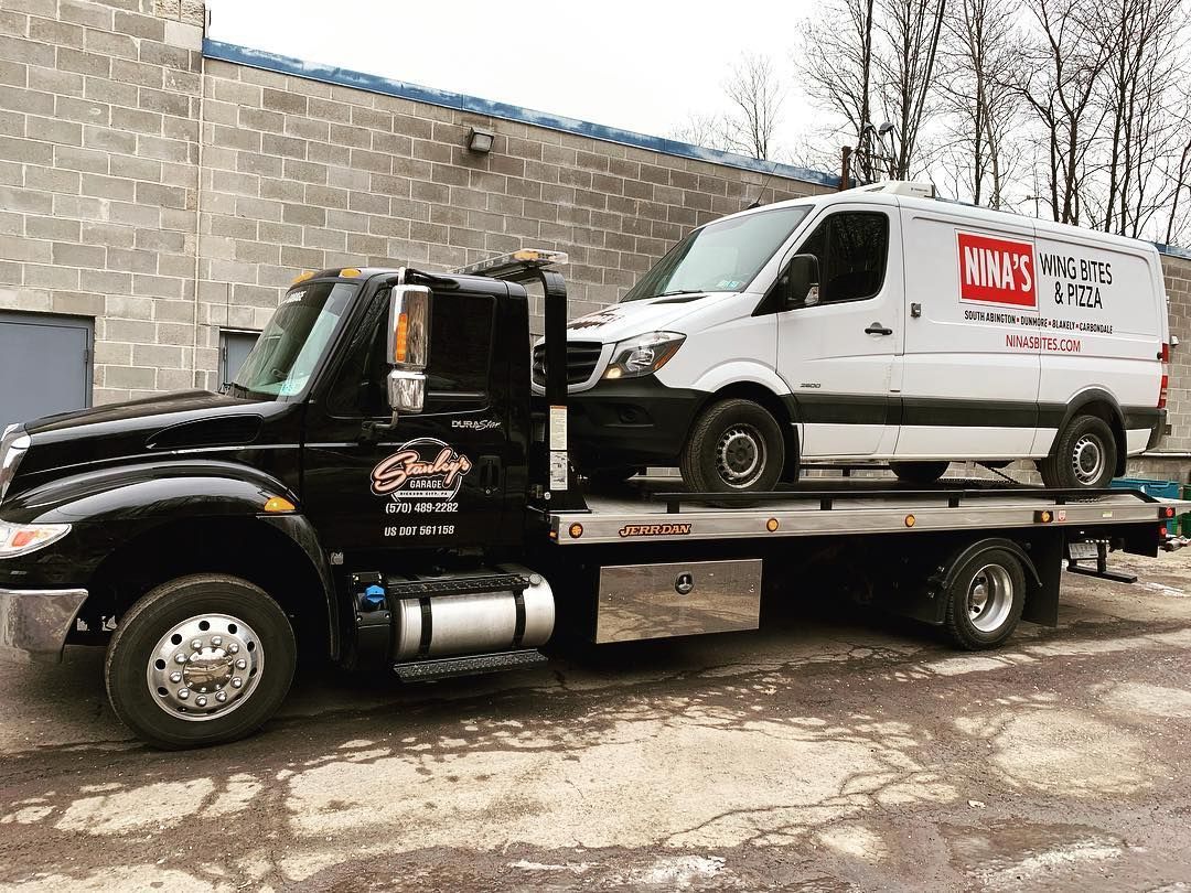 Black tow truck with a white van loaded on its flatbed. The van has business signage. Gray building background.