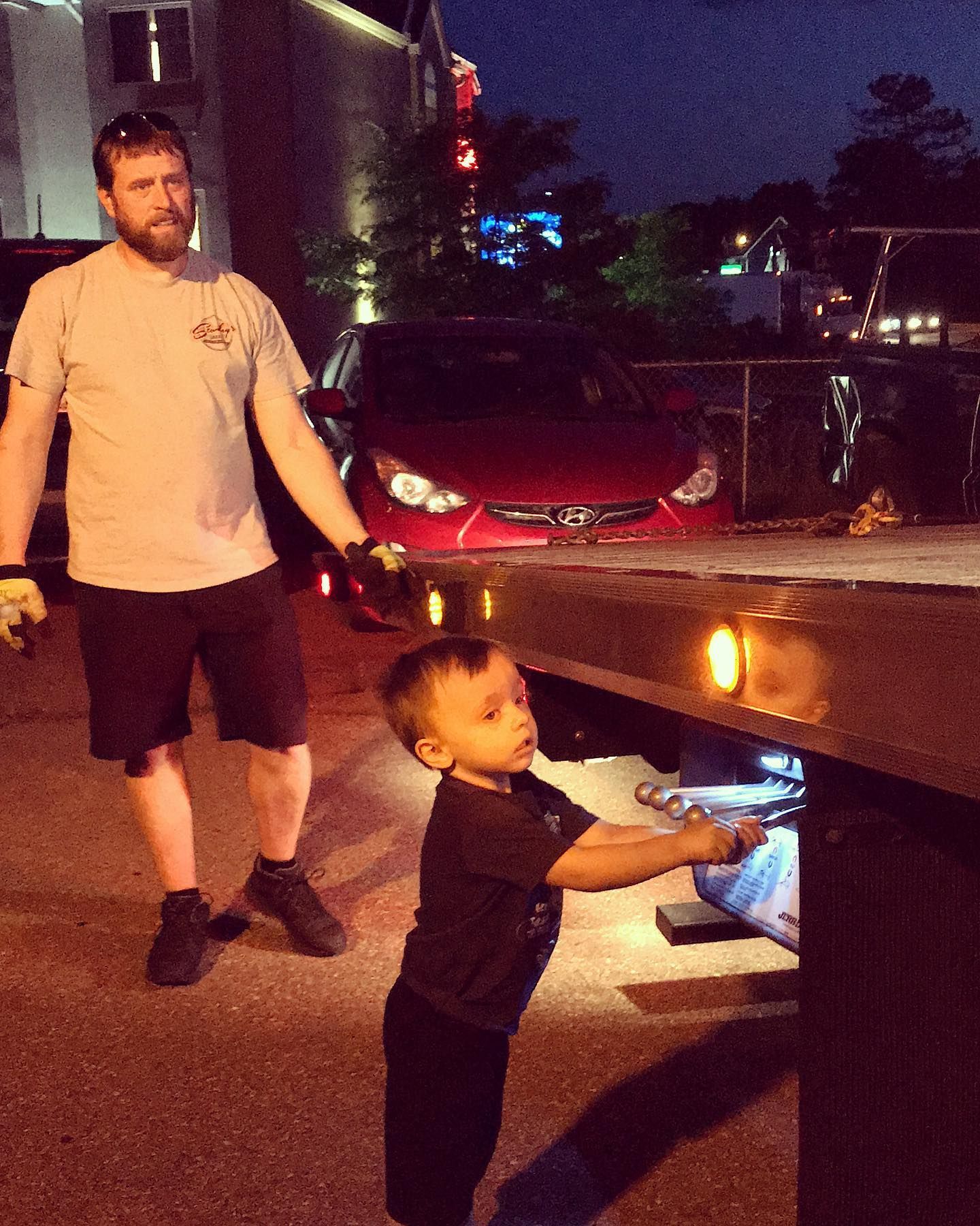 Man assists a young child examining the tow truck, red car in background at night.