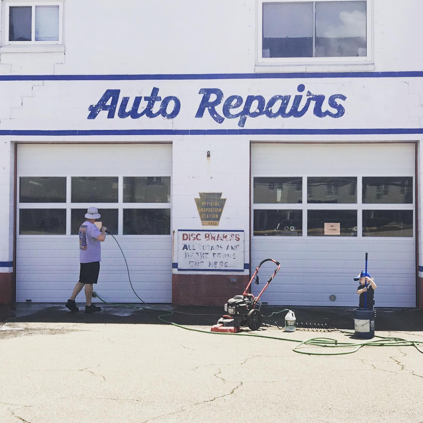 Two people cleaning the exterior of an auto repair shop with white garage doors, lawn mower in front.