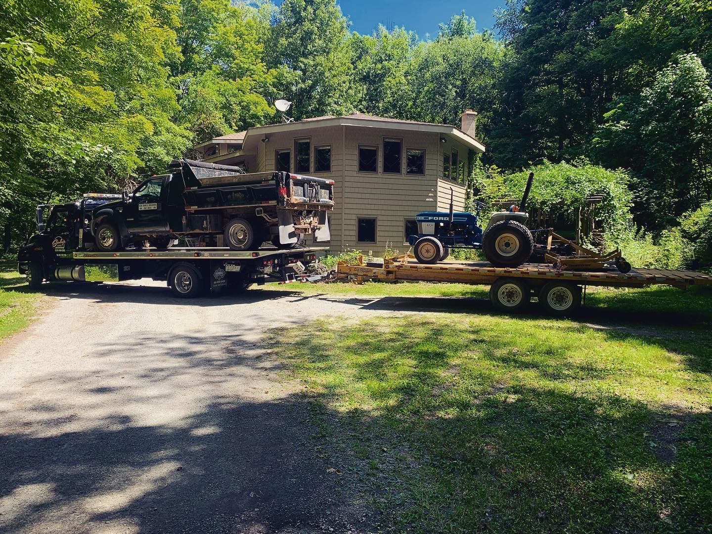 Two trucks with vehicles on trailers in front of a house, surrounded by trees.