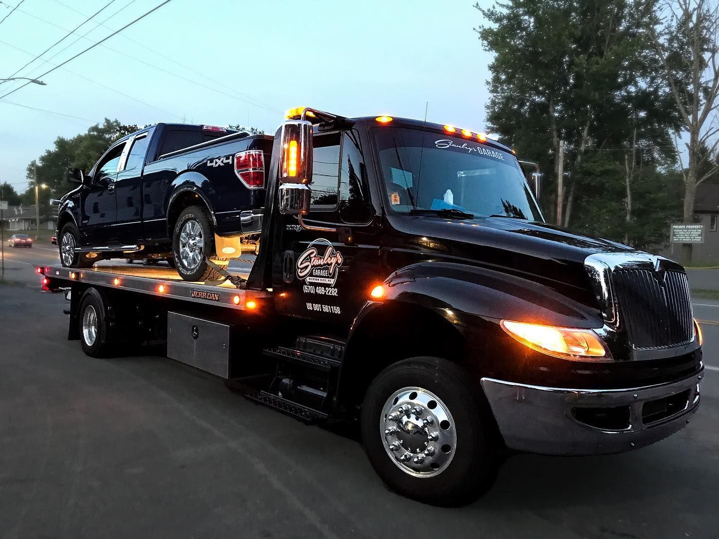 Black tow truck hauling a dark-colored pickup truck on a city street at dusk.