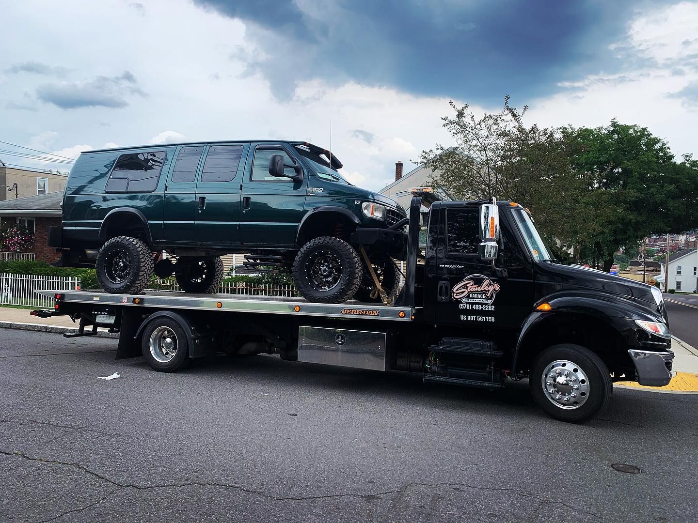 Green SUV on a flatbed tow truck. Black truck and SUV, asphalt road, overcast sky.