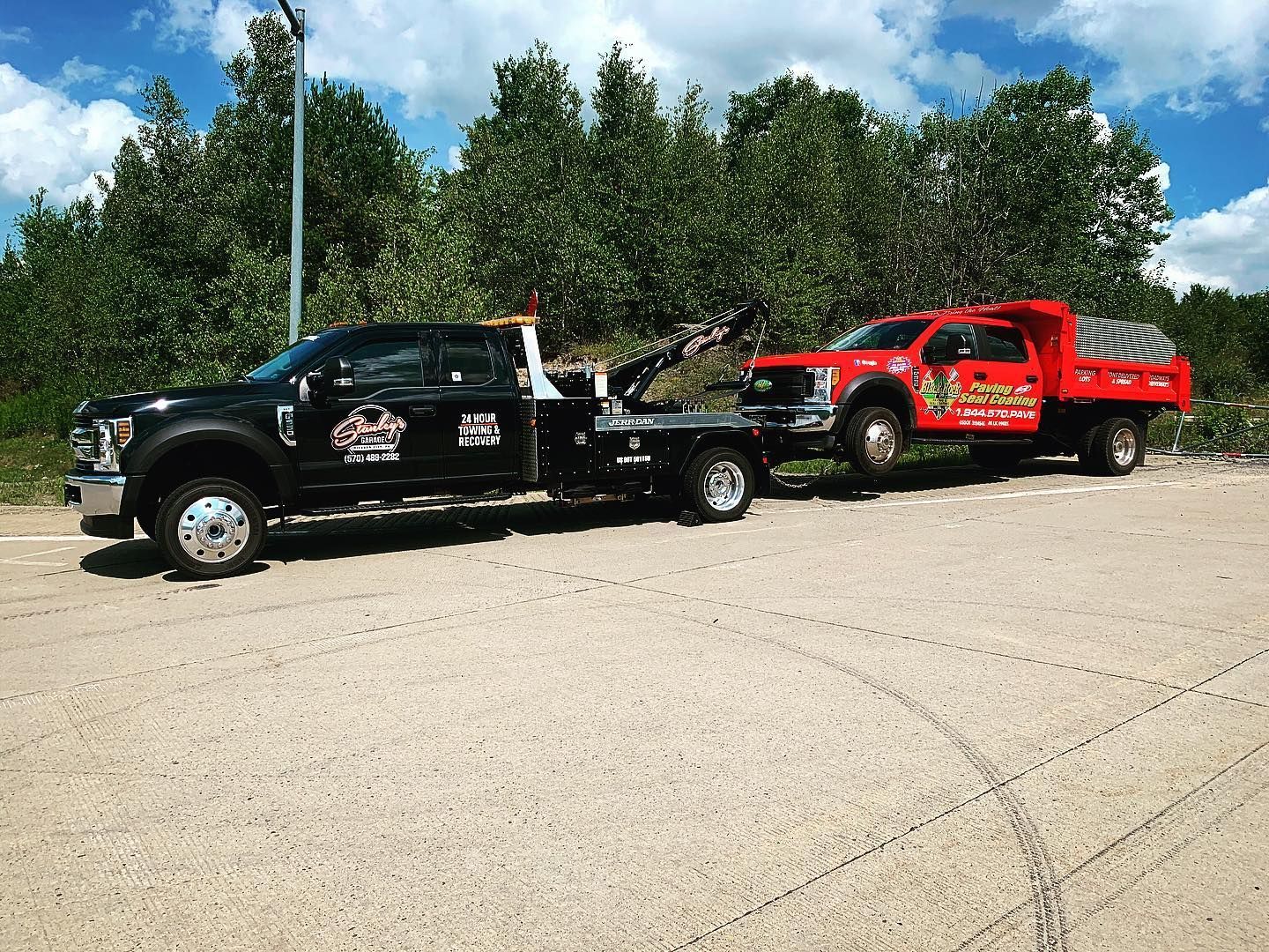 Black tow truck towing a red dump truck on a paved road; trees in background on a sunny day.